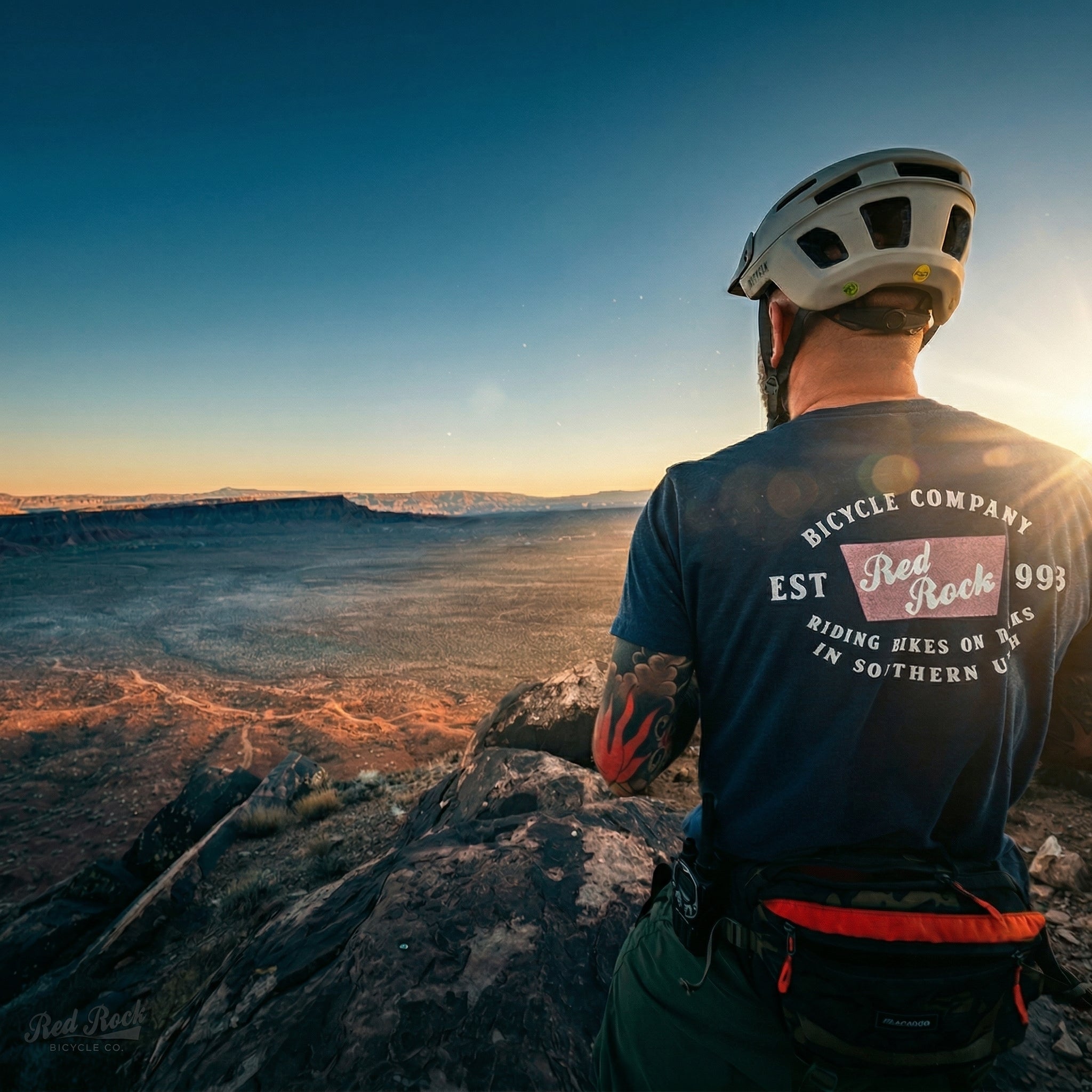Person wearing a helmet and bicycle company shirt overlooking a desert landscape at sunset.